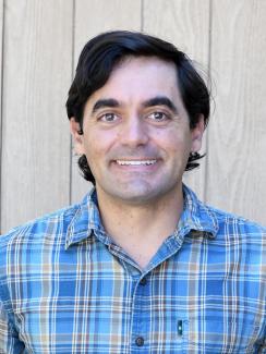Headshot of Jake Castellanos wearing a plaid button-down shirt with a fence behind him.